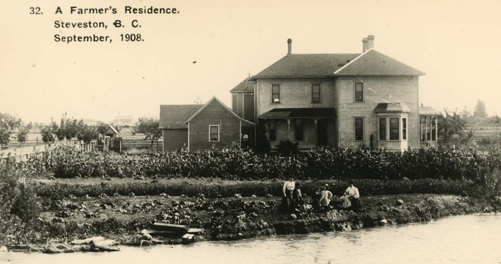 Sepia-coloured photo of a two-storey farmhouse behind a row of corn plants, and women and girls standing in the front, along a riverbank